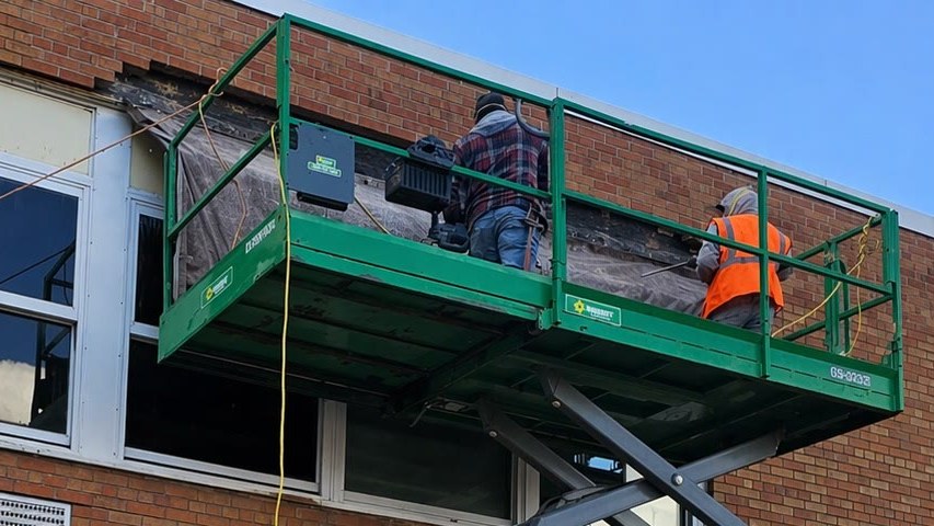 Masons removing deteriorated brick above a row of windows from a scissor lift