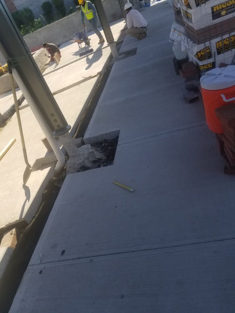 Newly poured concrete walkway sections with a cut-out for drainage and pallets of paving stones staged nearby