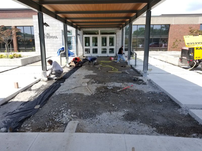 Crew preparing the sub-base at the Roslyn High School main entrance, with the canopy steel overhead and existing paving removed