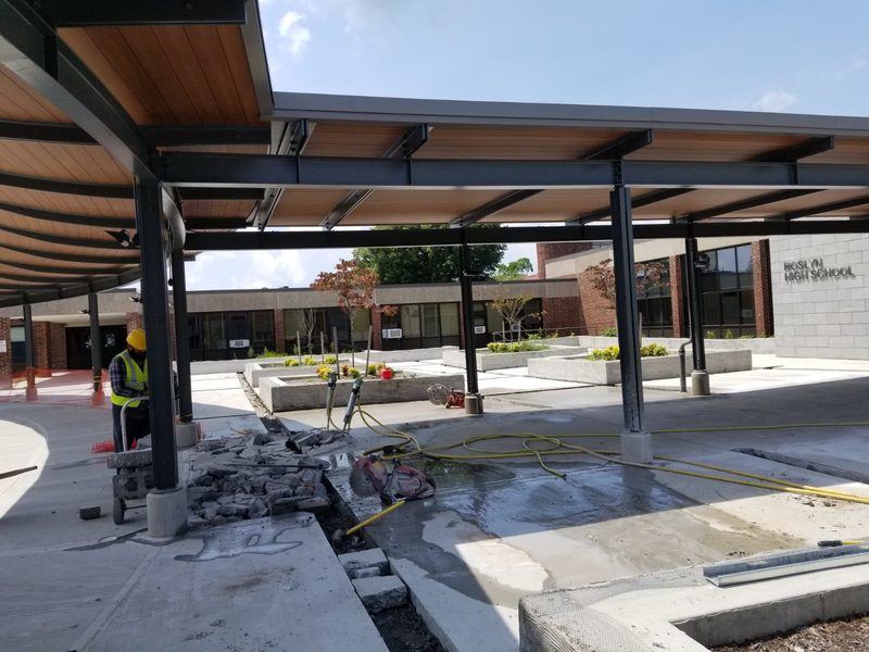 Worker saw-cutting concrete beneath the entrance canopy at Roslyn High School with completed concrete planters in the courtyard behind