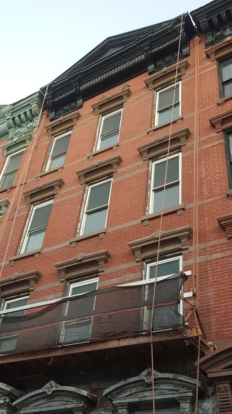 Suspended scaffold beneath the black mansard cornice of an ornate pre-war brick building with carved brownstone window pediments
